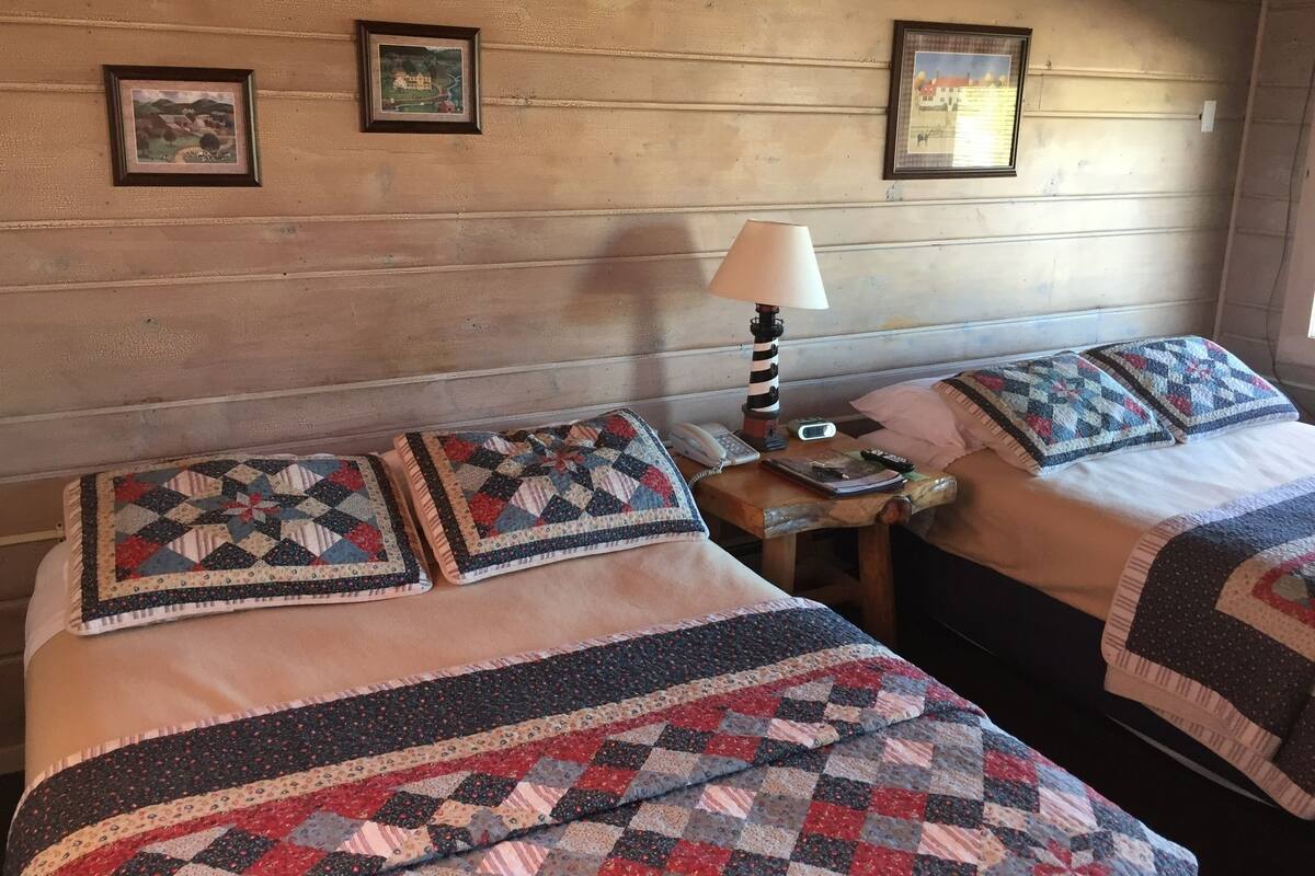 A room with two beds, each with a quilted bedspread and matching pillows. There is a nightstand with a lamp, a phone, and a remote control. Two framed pictures are hung on the wall above the beds.