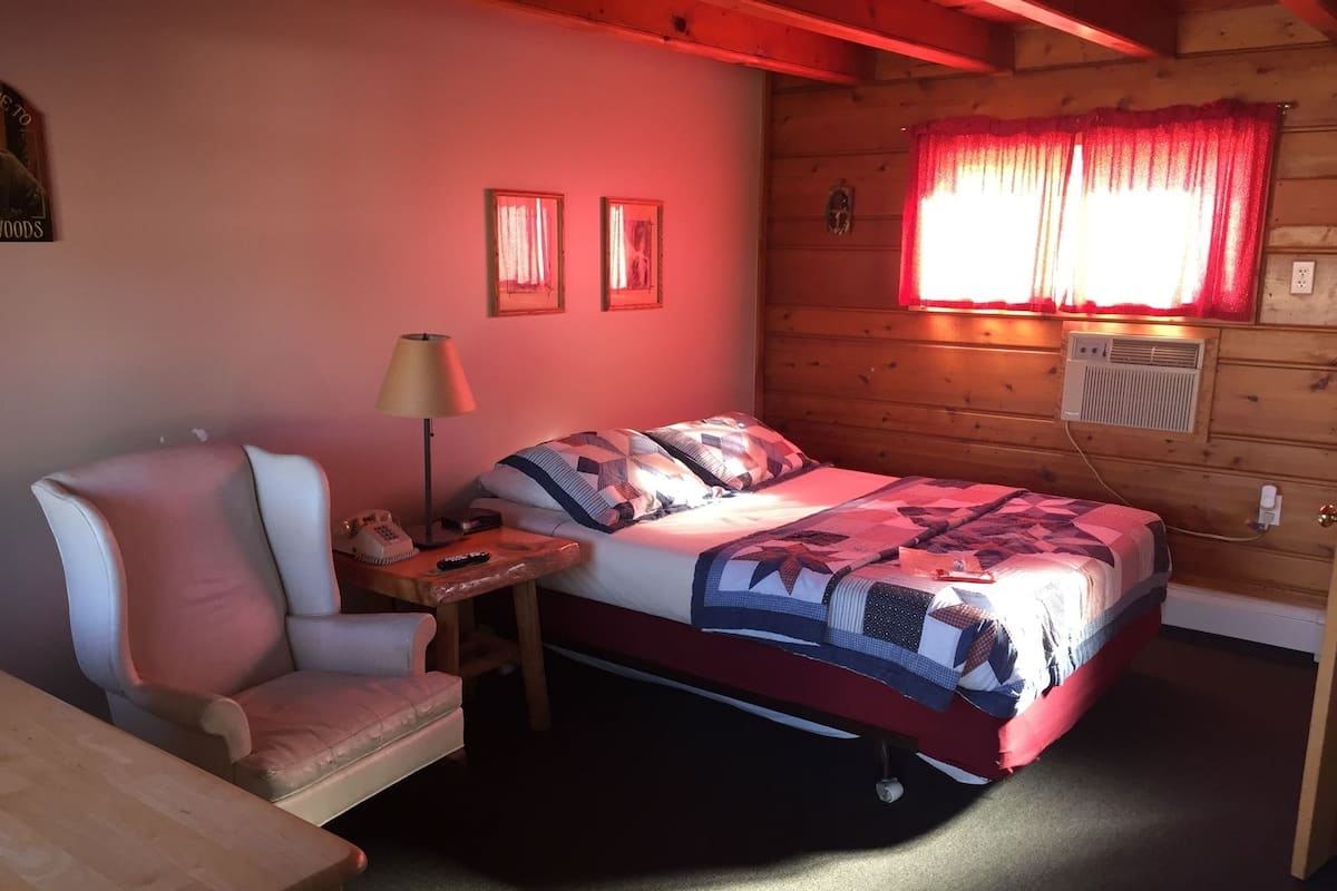 A cozy room with a bed, a chair, a small table with a lamp, and a window with curtains.