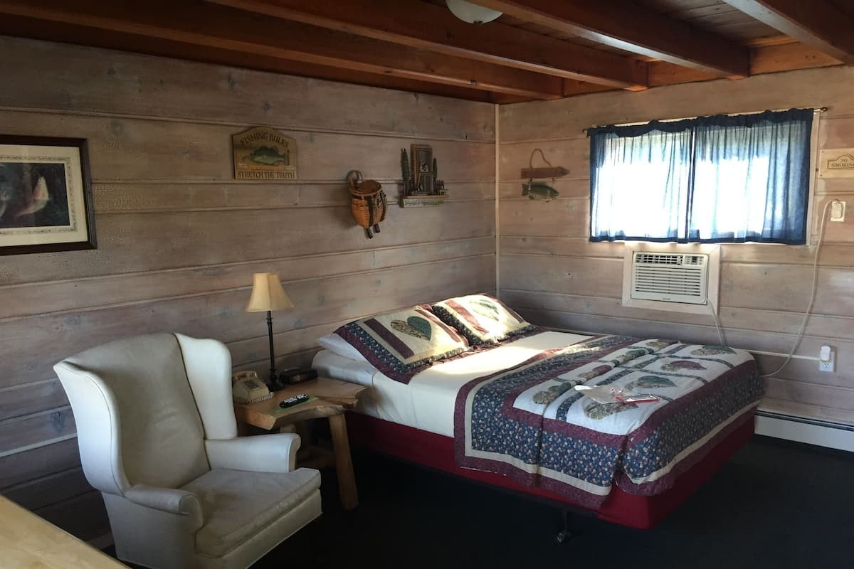 A cozy cabin-style room with a bed, a chair, a nightstand, and a lamp.