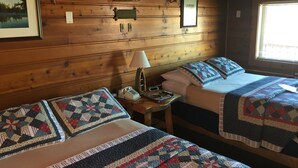A cozy cabin room with two beds, a wooden wall, a framed picture, a lamp, and a window.