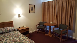 Une chambre d’hôtel avec un lit, un bureau avec un téléphone, deux chaises autour d’une petite table, une lampe et une fenêtre avec des rideaux.