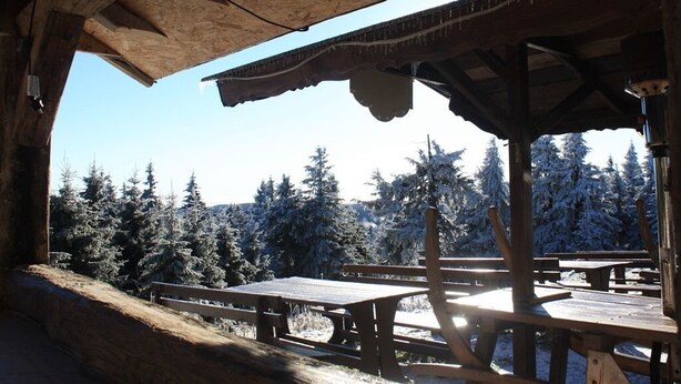 A wooden cabin with a covered outdoor area, featuring picnic tables and a view of snow-covered trees. - Fichtelberghütte (Oberwiesenthal)