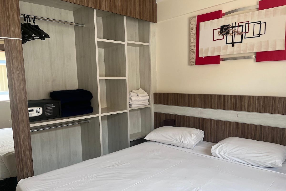 A hotel room with a bed, a control panel, a shelf with towels, and a wall art piece.