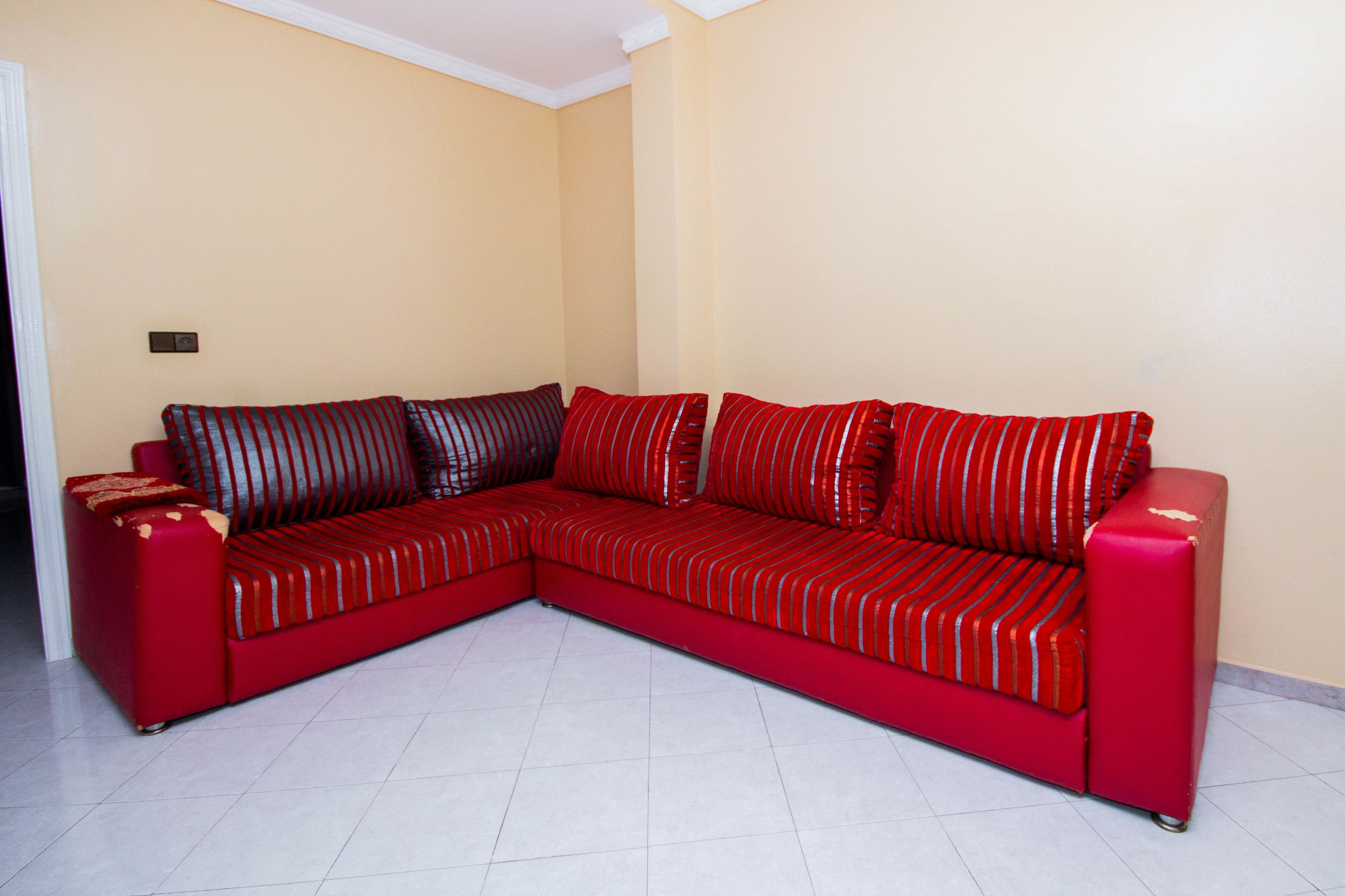 Un canapé d’angle rouge avec des coussins à rayures, dans une pièce aux murs de couleur beige et au sol carrelé de carreaux blancs.