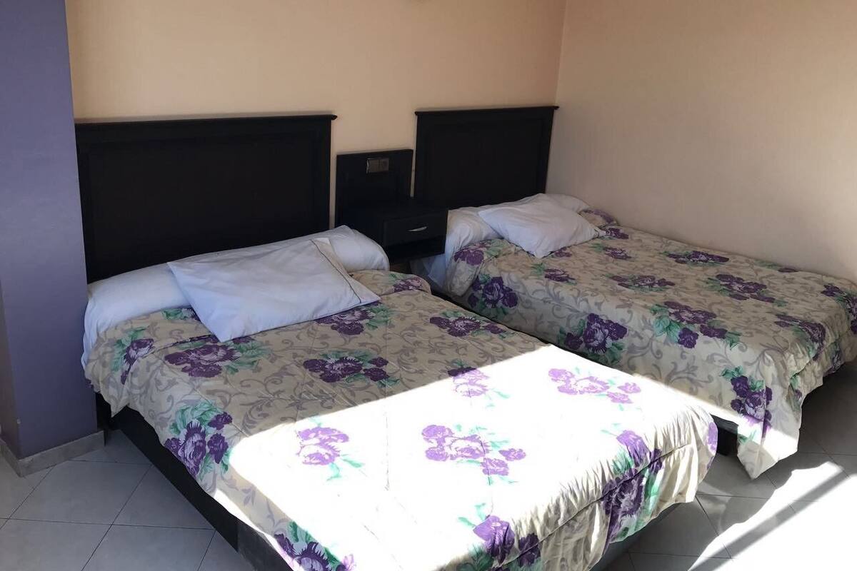 A room with two beds, each with floral bedding and white pillows. There is a small bedside table with a lamp between the beds.