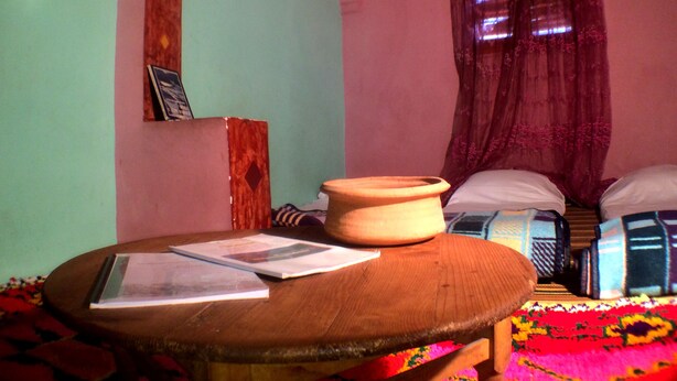 A room with a bed, a table with papers, a wooden stool, a colorful rug, and a wall with a mirror and a picture frame. - Atlas Kasbah Lodge (Asni)