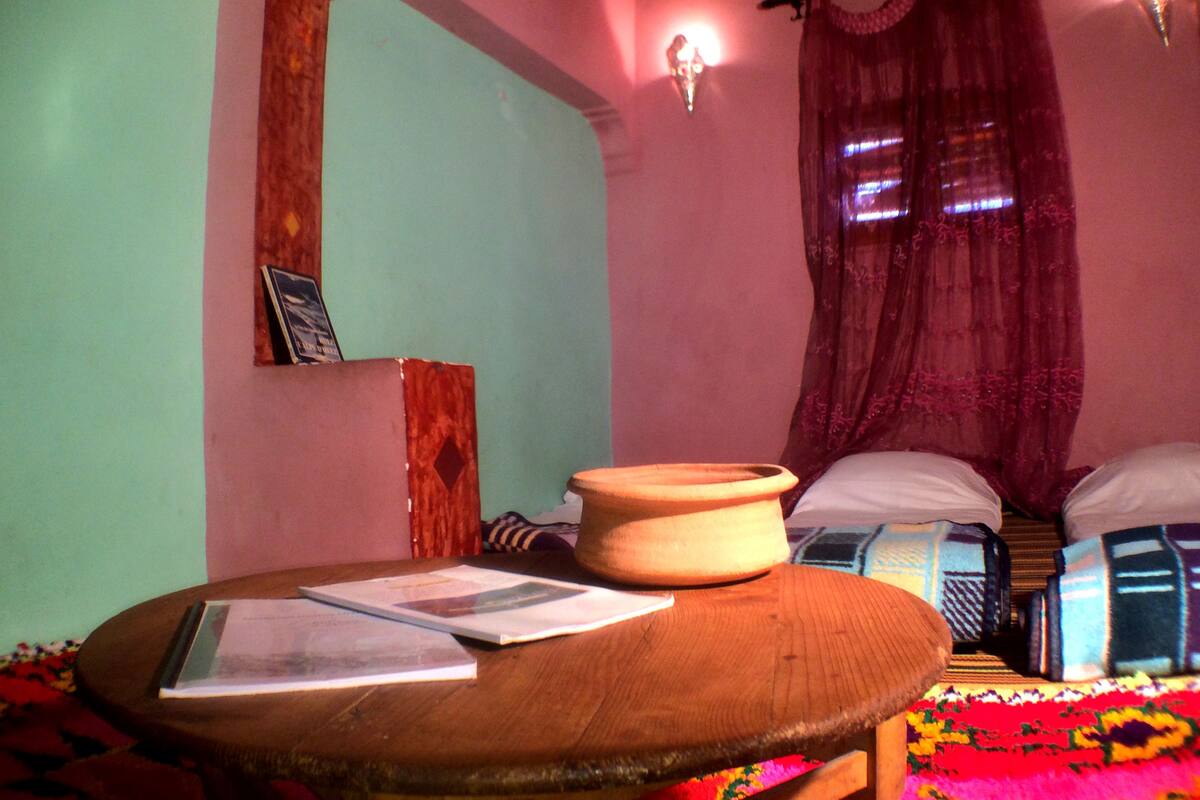 A room with a bed, a table with papers, a wooden stool, a colorful rug, and a wall with a mirror and a picture frame.