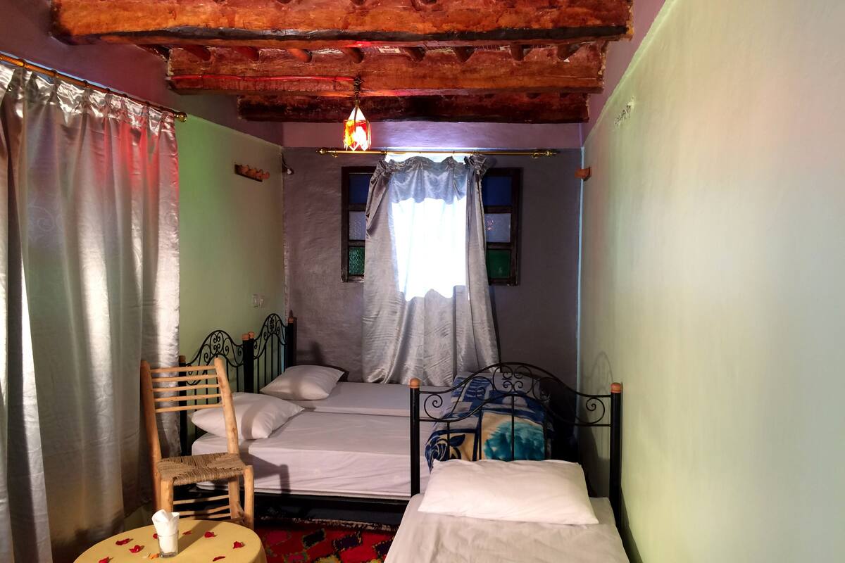 A small, cozy bedroom with two beds, a wooden chair, a small round table with a napkin, and a colorful rug.