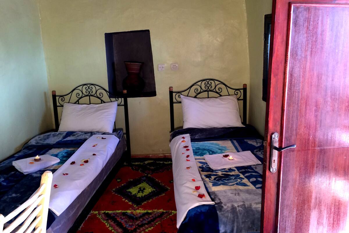 Two single beds with white pillows and blue bedding, a small table with a candle, and a red door.