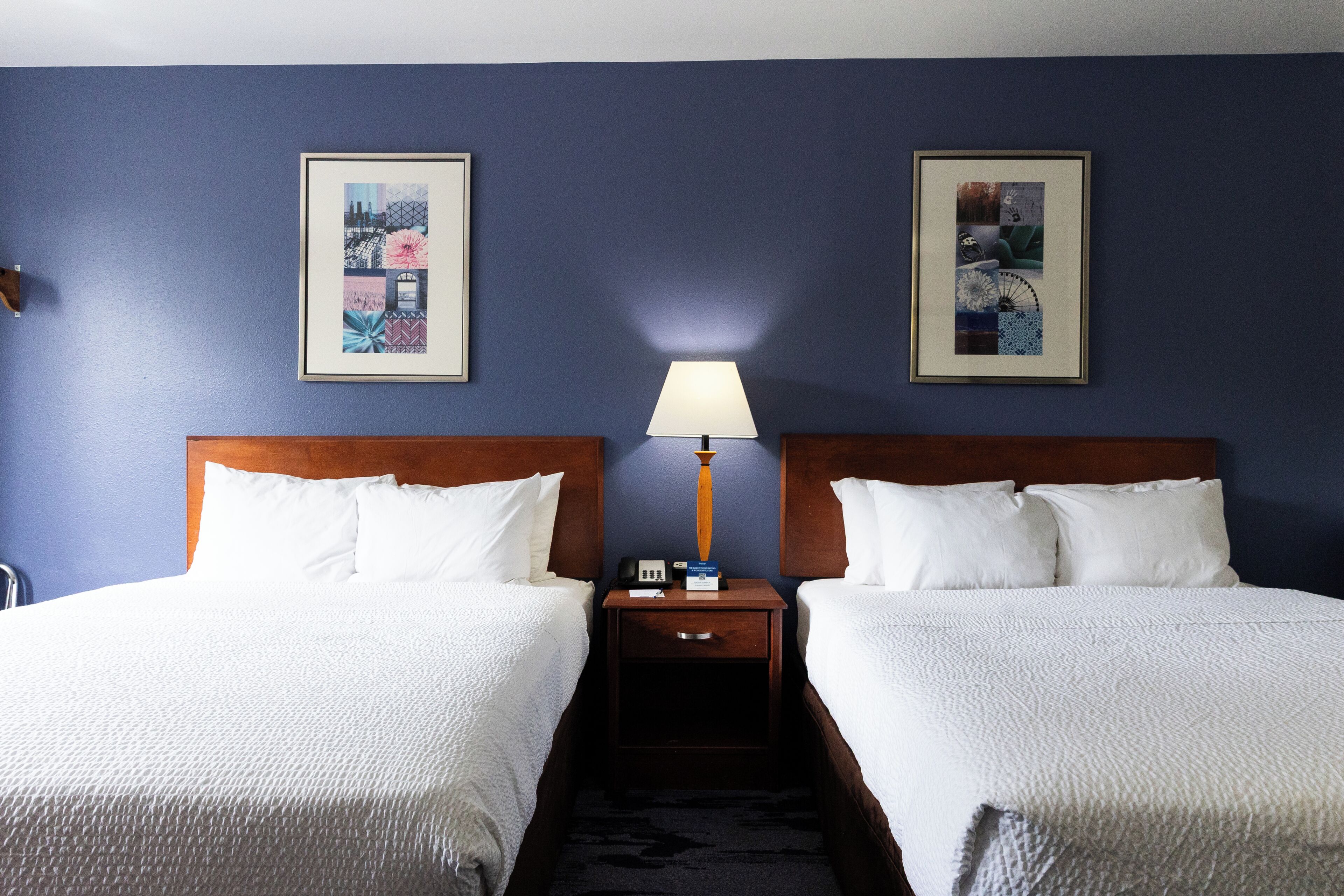 Una habitación de hotel con dos camas, una mesita de noche con lámpara y dos cuadros en la pared.