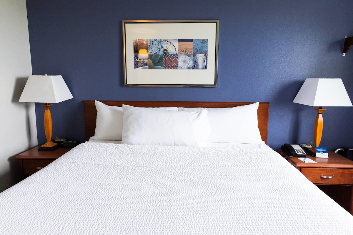 A hotel room with a large bed, two bedside tables, a framed artwork on the wall, and a nightstand with a lamp.