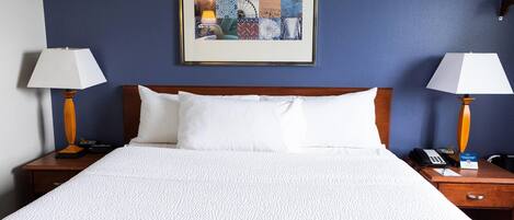 A hotel room with a large bed, two bedside tables, a framed artwork on the wall, and a nightstand with a lamp.
