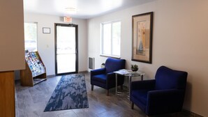A lobby with a reception desk, a blue armchair, a small table with a plant, a framed picture on the wall, and a carpeted floor.