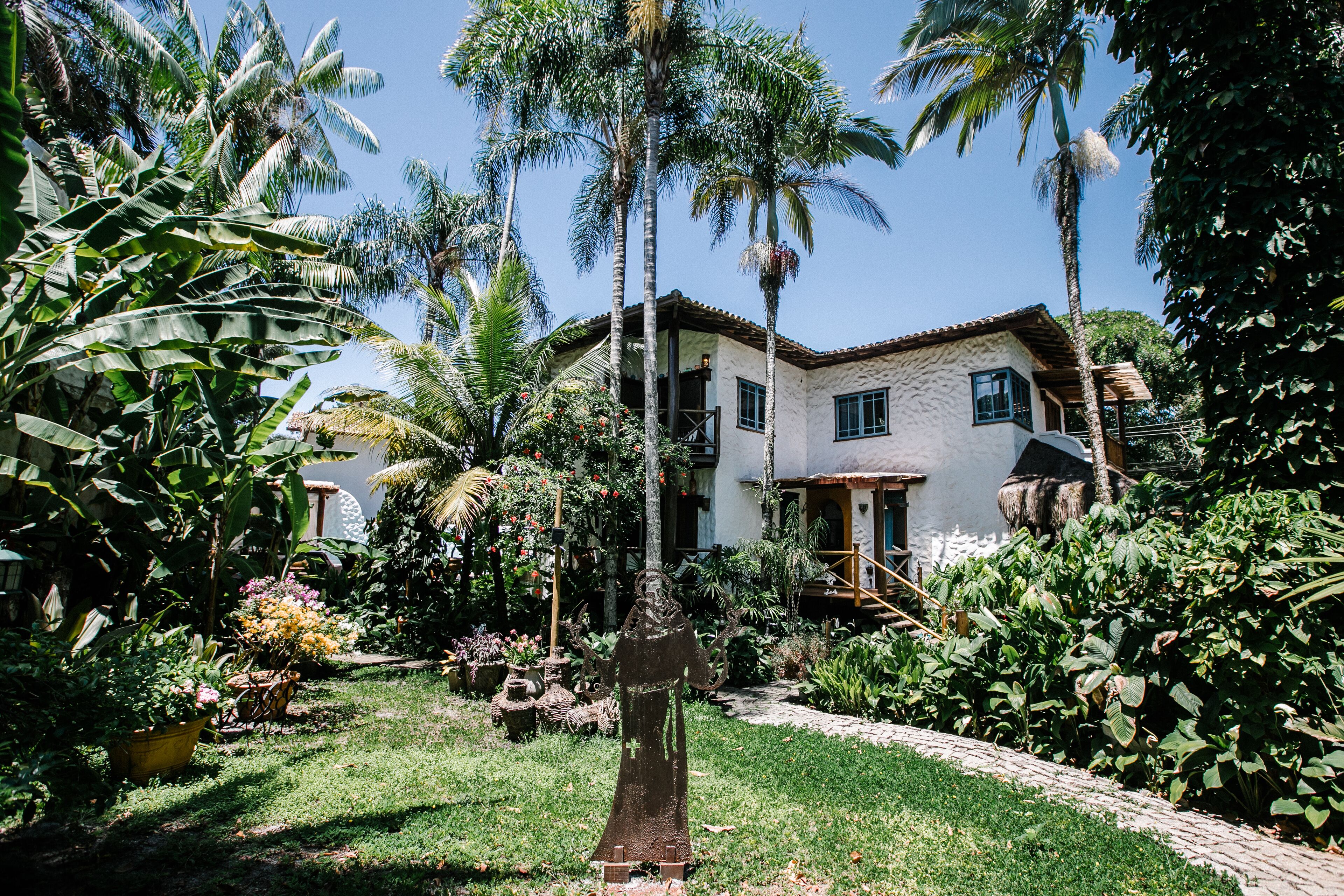 A two-story house with a garden, surrounded by palm trees and various plants.