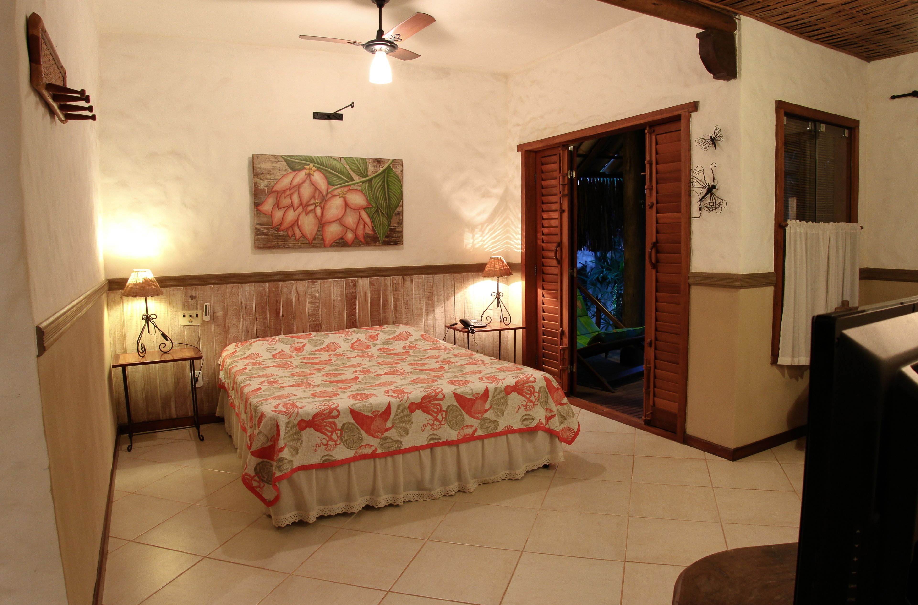 A bedroom with a bed, two bedside lamps, a ceiling fan, a painting on the wall, and a window with shutters.