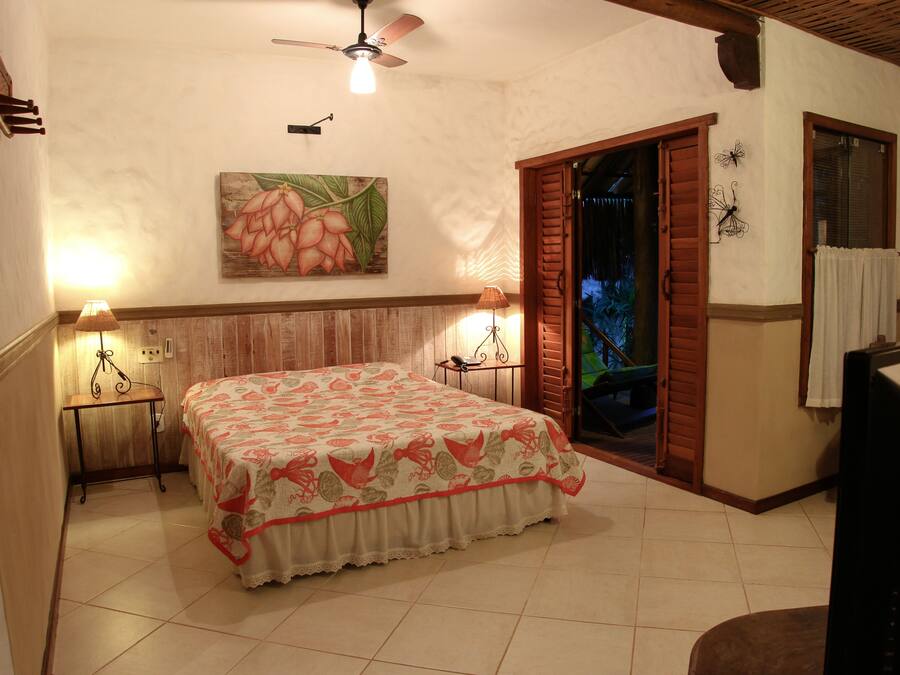 A bedroom with a bed, two bedside lamps, a ceiling fan, a painting on the wall, and a window with shutters.