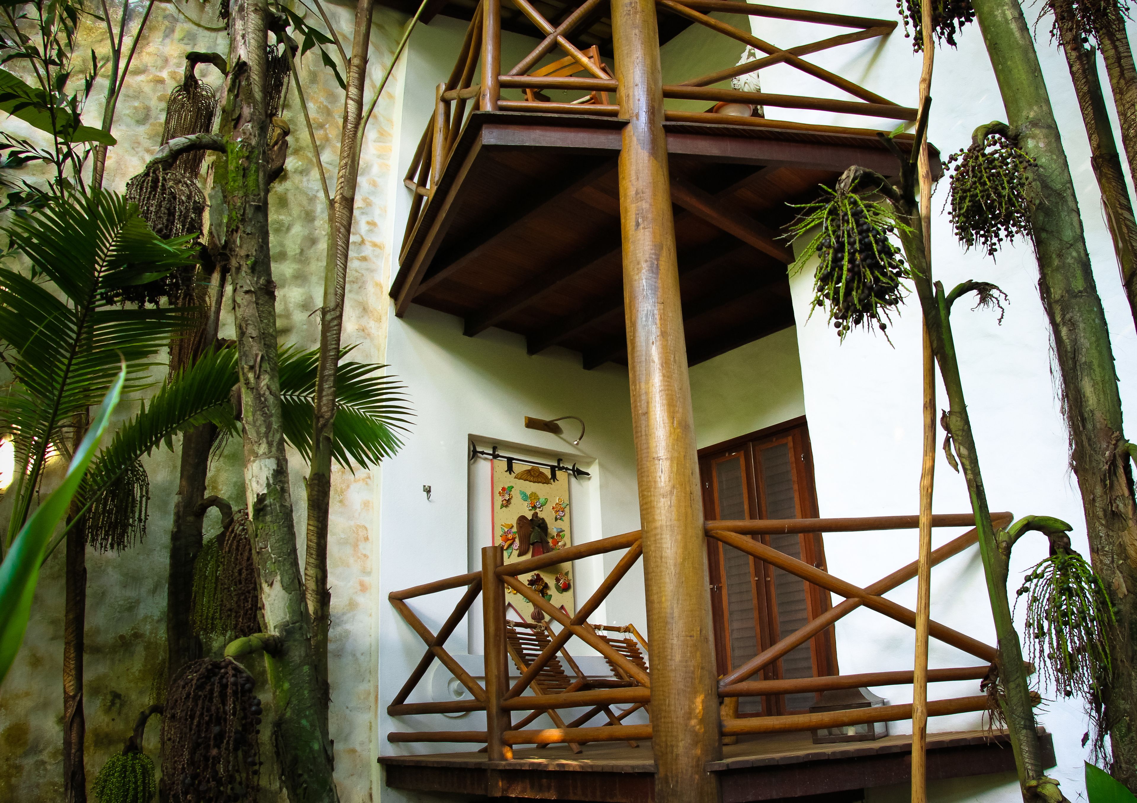 A wooden balcony with a view of a building's interior, surrounded by tropical plants and a rustic wooden railing.