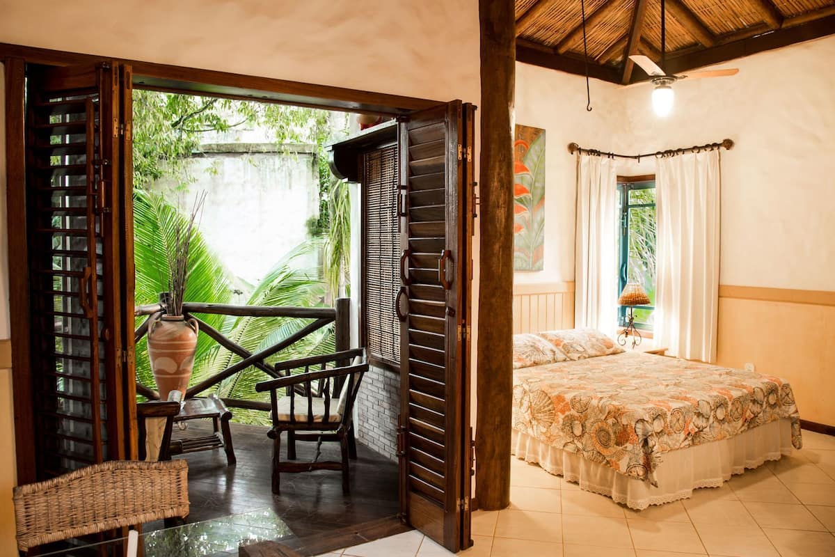 Una habitación con cama, ventana con cortinas, ventilador de techo y vistas al exterior.