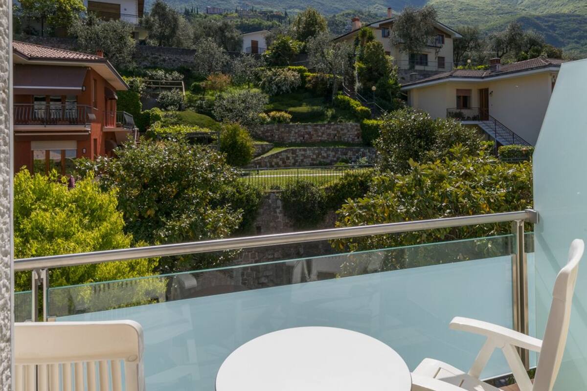 A balcony with a glass railing, a round table, and two white chairs. In the background, there are houses, trees, and mountains.