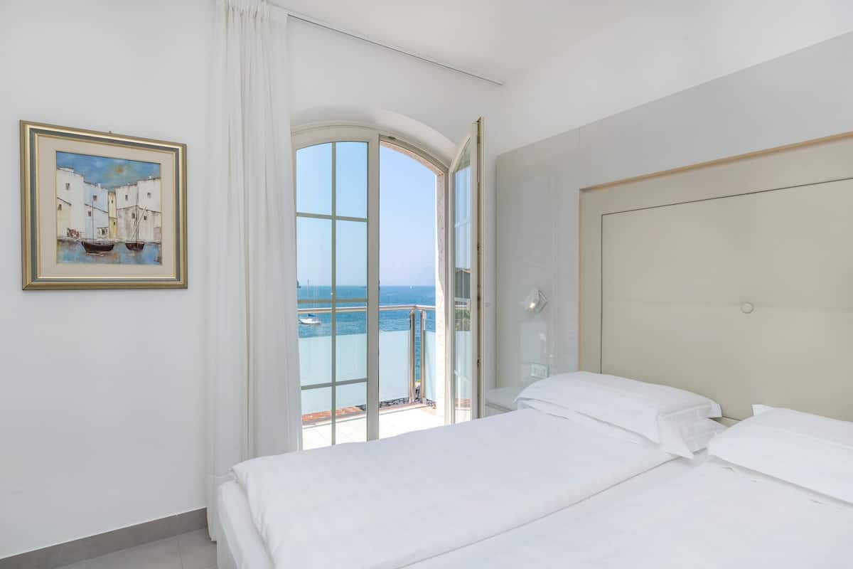 A hotel room with a bed, a painting, and a balcony with a view of the sea.