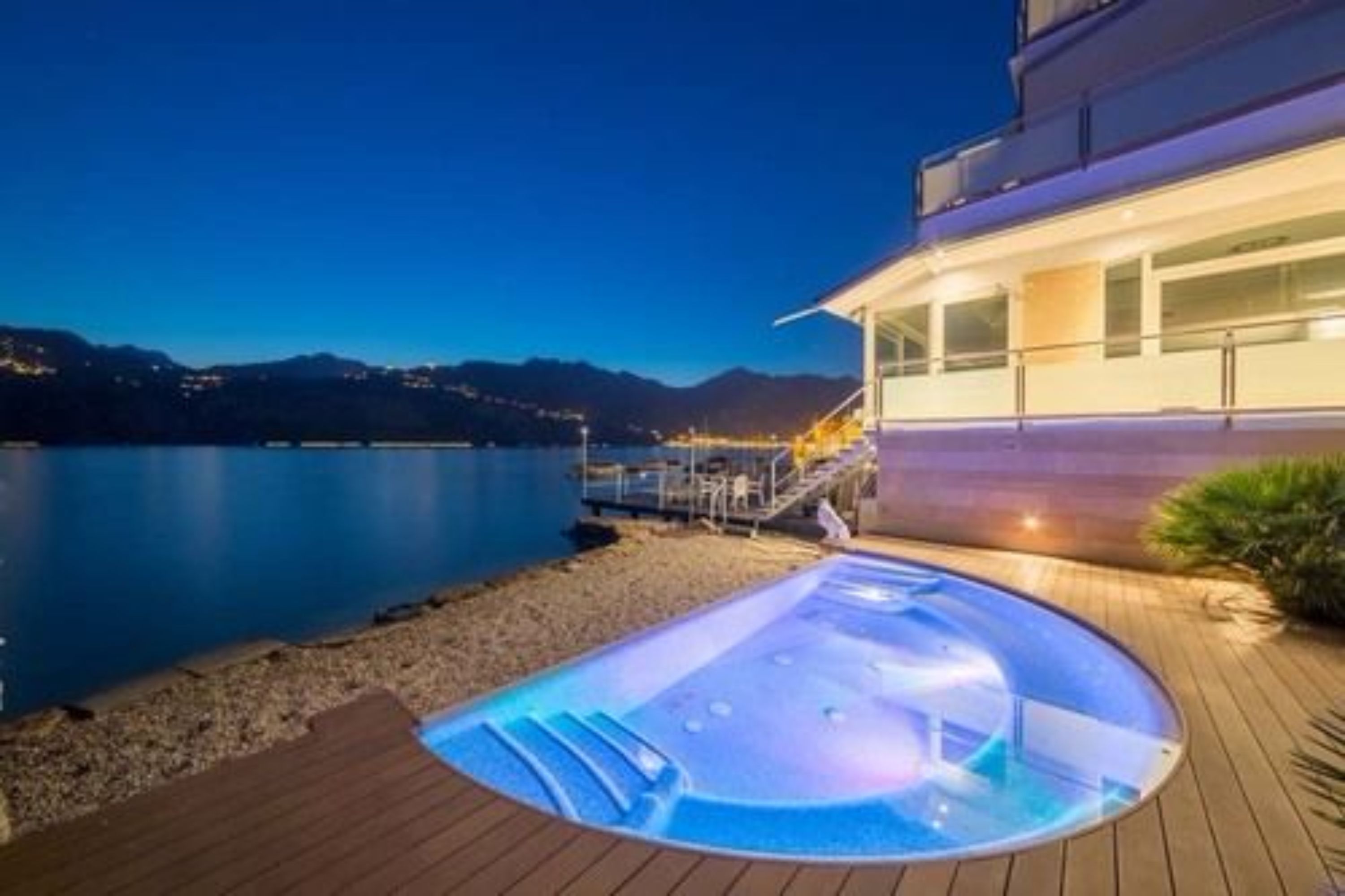 A modern building with a hot tub and a wooden deck by a lake at night.