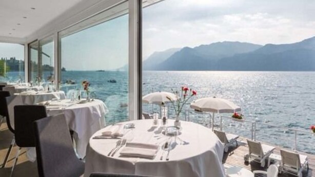 Un restaurant où les tables sont dressées pour le service, avec de grandes fenêtres offrant une vue sur la mer et les montagnes.