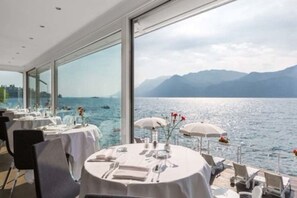 A restaurant with tables set for dining, large windows offering a view of the sea and mountains. - Hotel Vega (Malcesine)