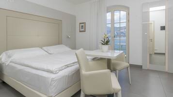 A hotel room with a bed, a desk, a chair, a potted plant, and a view of the sea.
