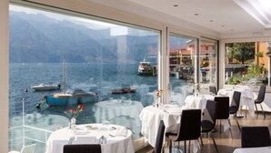 Un restaurant avec des tables dressées pour le dîner, de grandes fenêtres donnant sur un lac où l’on peut admirer les bateaux, et une chaîne de montagnes en toile de fond.