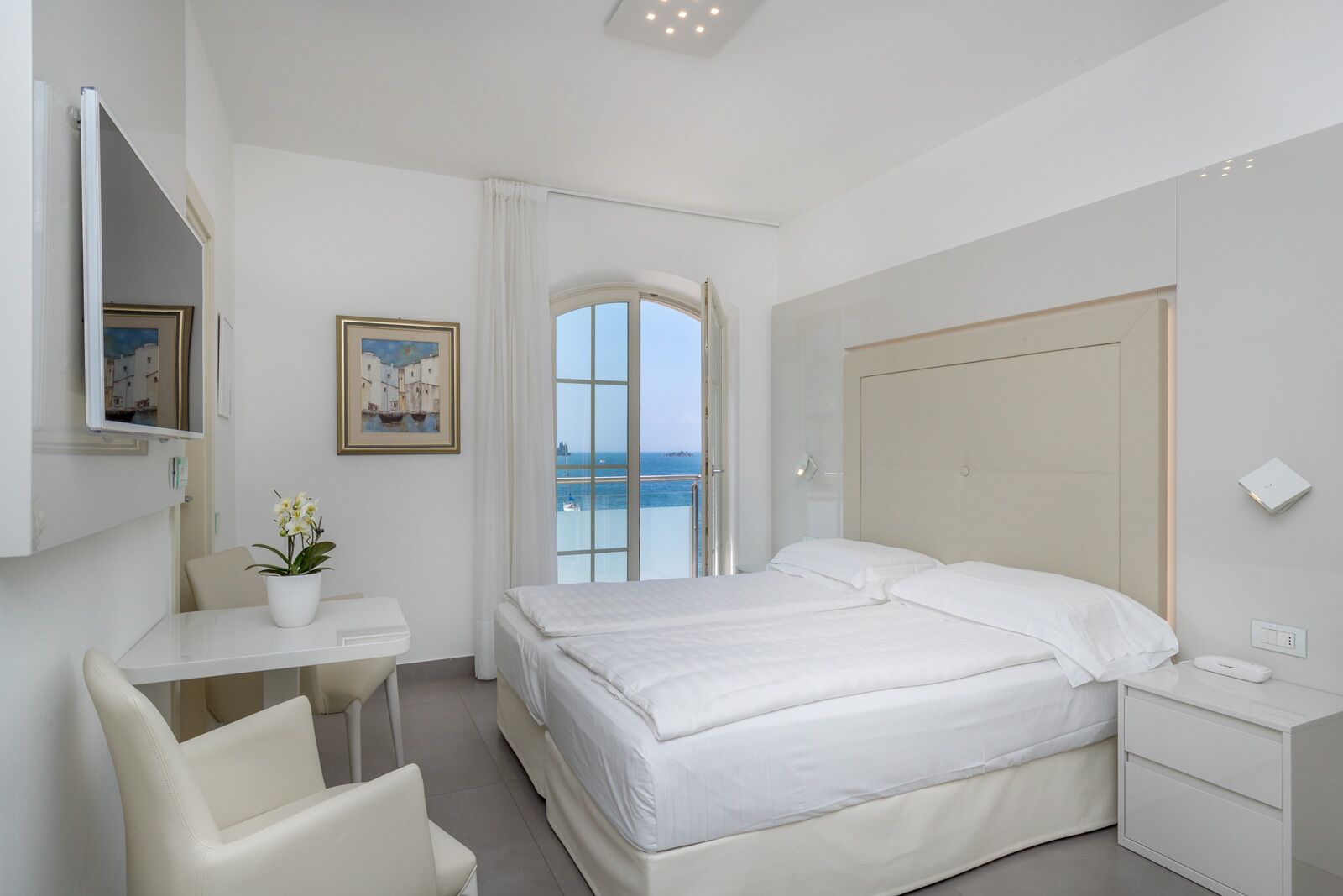 A hotel room with a bed, a desk, a chair, a small table with a potted plant, and a view of the sea through a window.