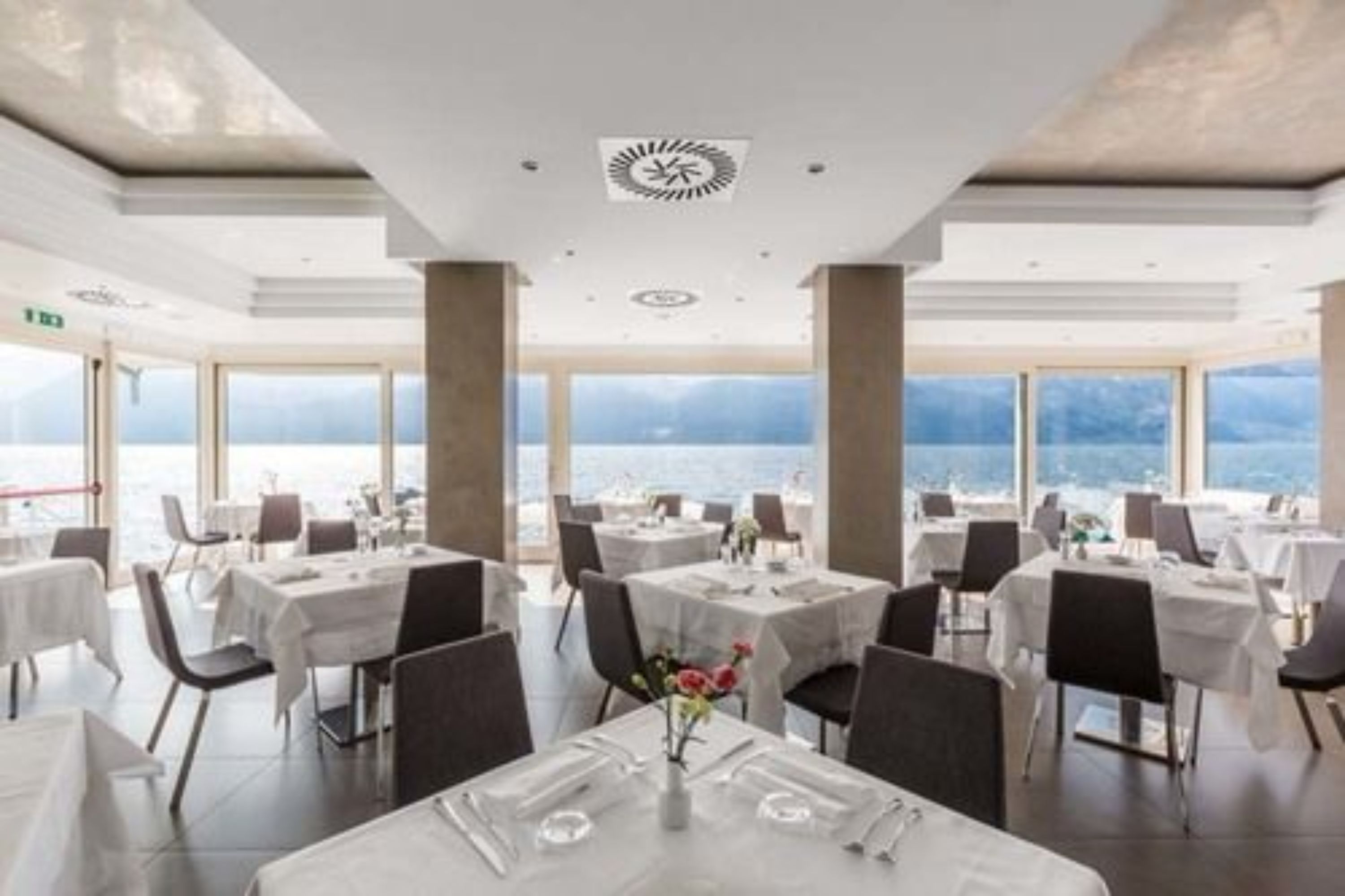 A restaurant with tables set for dining, large windows offering a view of the sea, and a modern ceiling design.