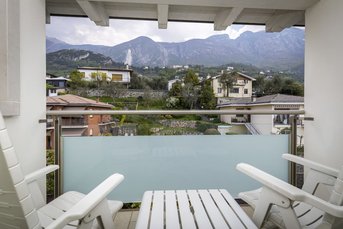 A balcony with white chairs and a table, overlooking a mountainous landscape with houses and greenery.