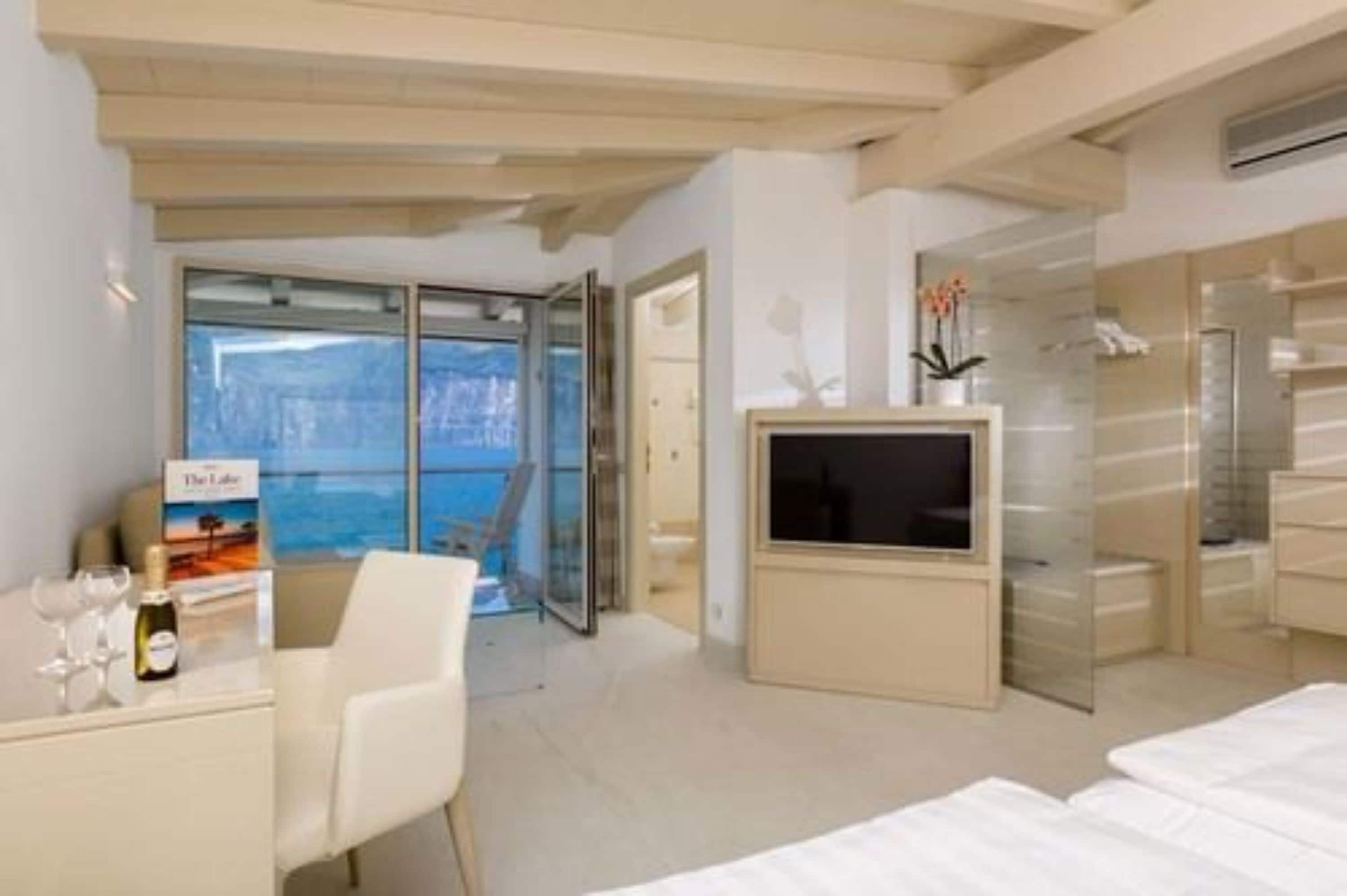 A modern hotel room with a large window, a flat-screen TV, a minibar, and a dining area with a table and chairs.