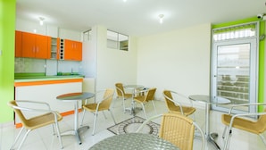 A modern kitchen with orange cabinets, a dining area with wicker chairs and round tables, and a glass door with a security camera. - Hostal Florida (Chiclayo)