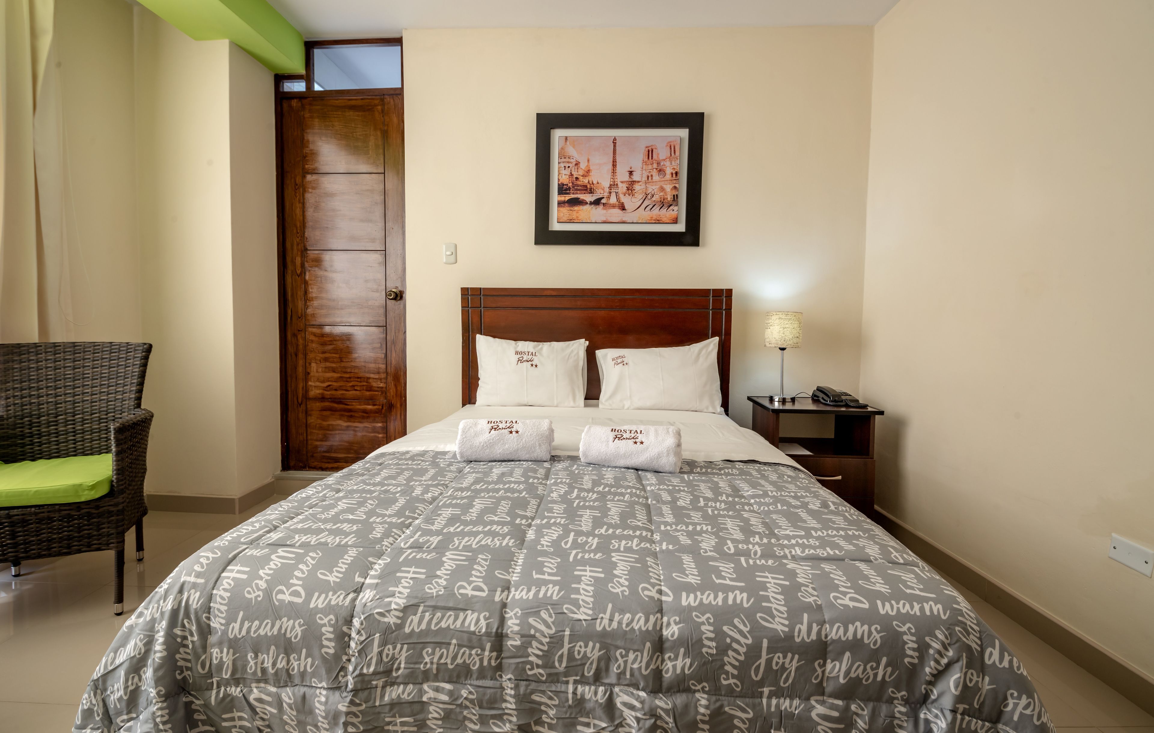 Habitación de hotel ordenada con cama, una silla, una mesita de noche y un cuadro enmarcado con una vista urbana en la pared.