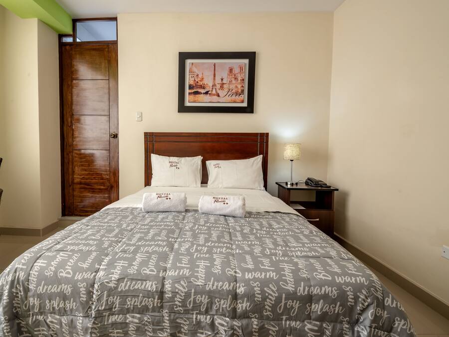 Quarto de hotel bem arrumado, com uma cama, uma cadeira, uma mesa de cabeceira e um quadro com uma vista urbana na parede.