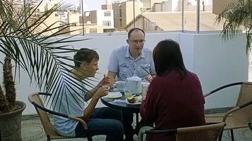 Un grupo de personas cenando en una mesa al aire libre con vistas a la ciudad, rodeado de macetas con plantas.