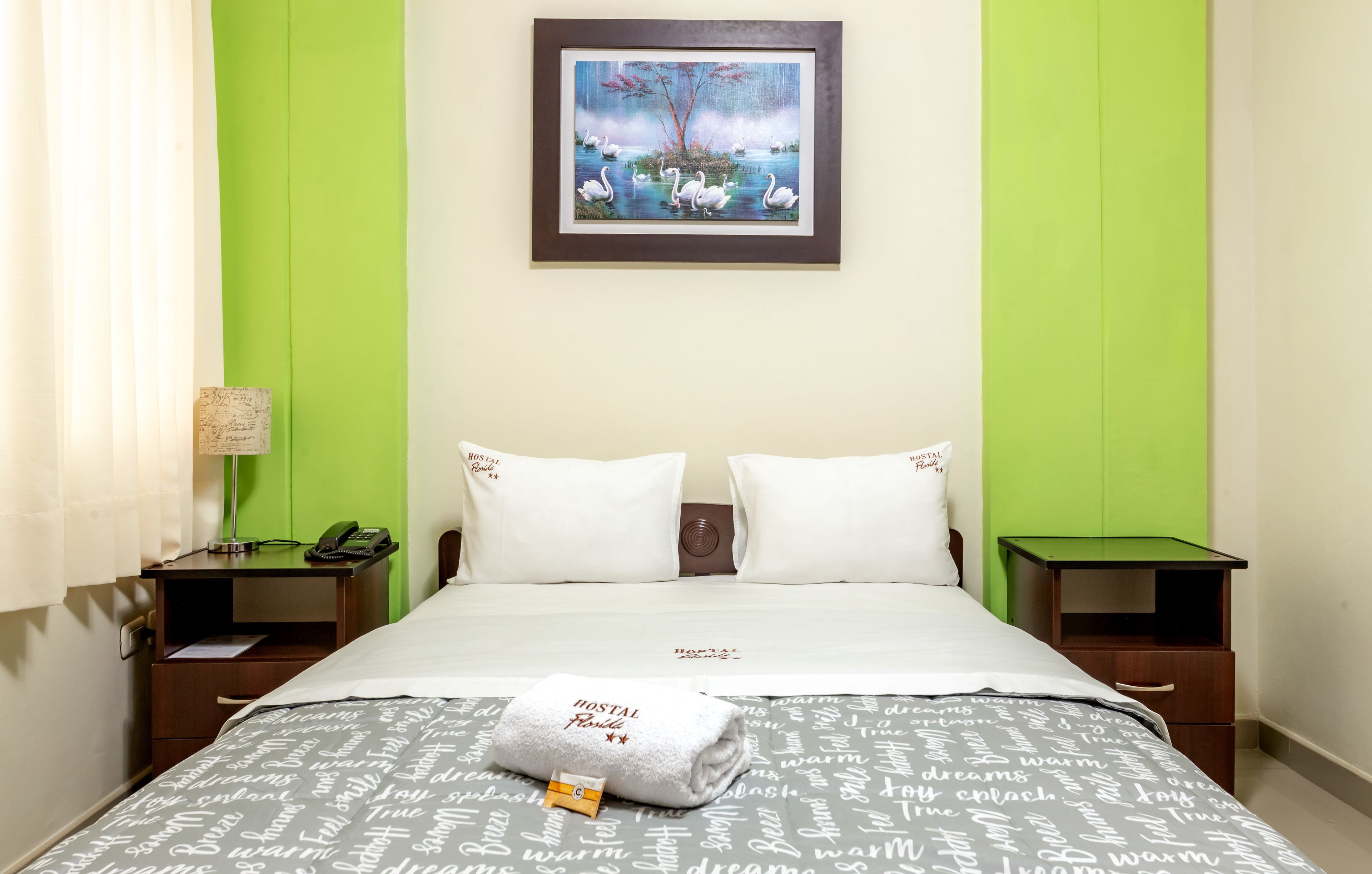 Habitación de hotel con cama, dos mesitas de noche, lámpara, un cuadro y pared verde.