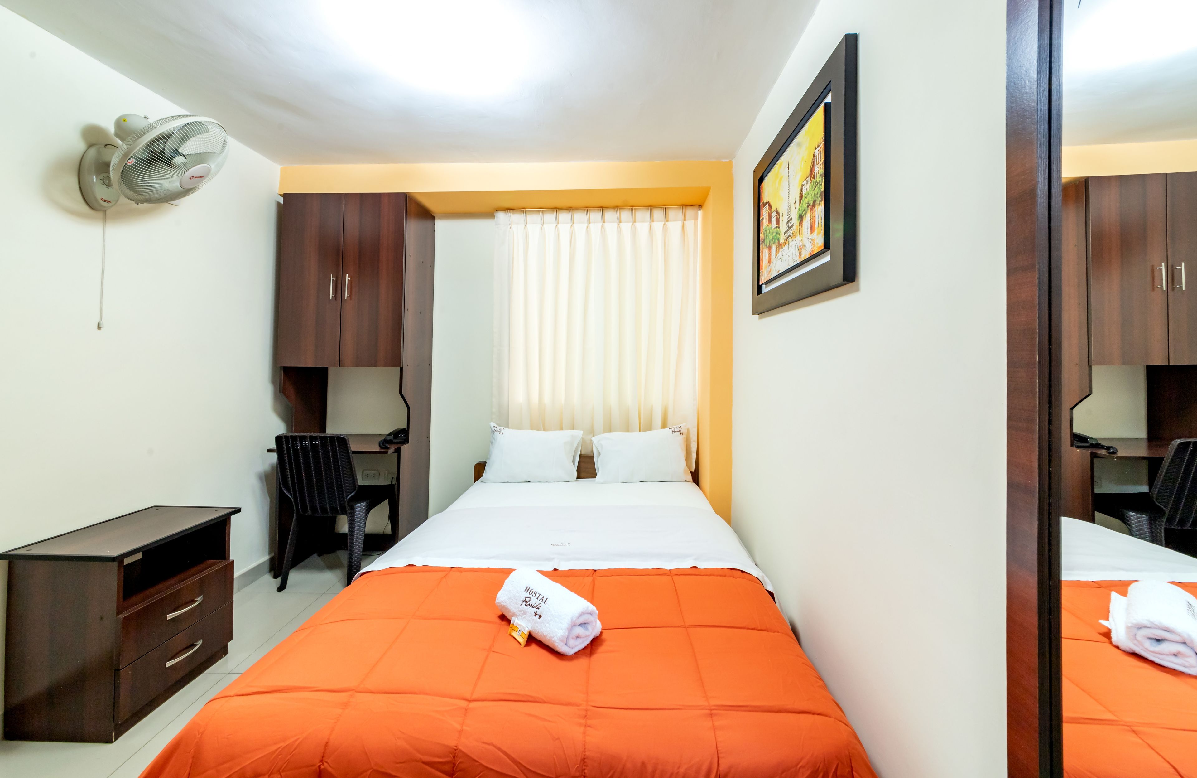 A hotel room with a bed, a desk, a chair, a wardrobe, and a ceiling fan.