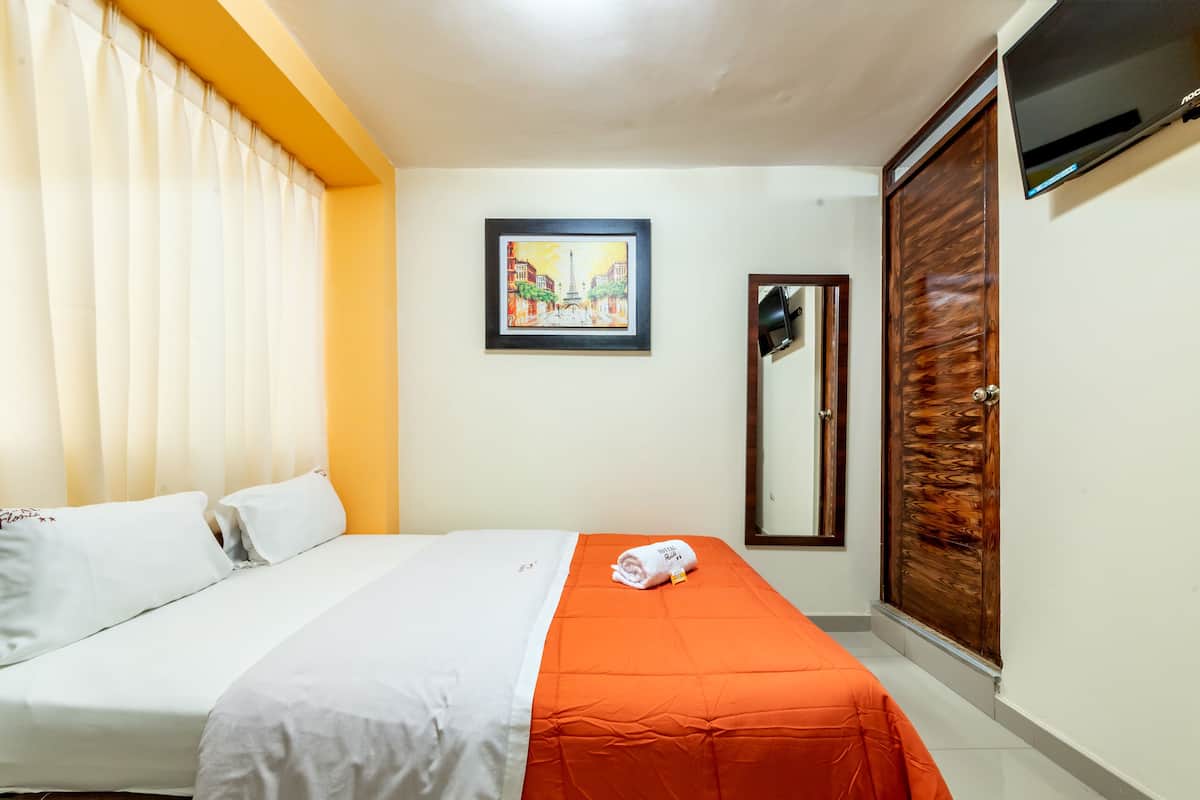 A hotel room with a bed, a wooden wardrobe, a framed picture on the wall, and a TV mounted above the wardrobe.