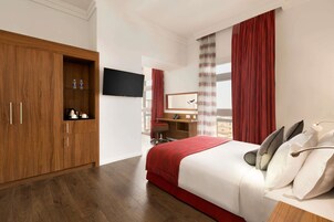 Habitación de hotel con un armario de madera, una cama con ropa de cama blanca y roja, un escritorio con una silla y un televisor montado en la pared.
