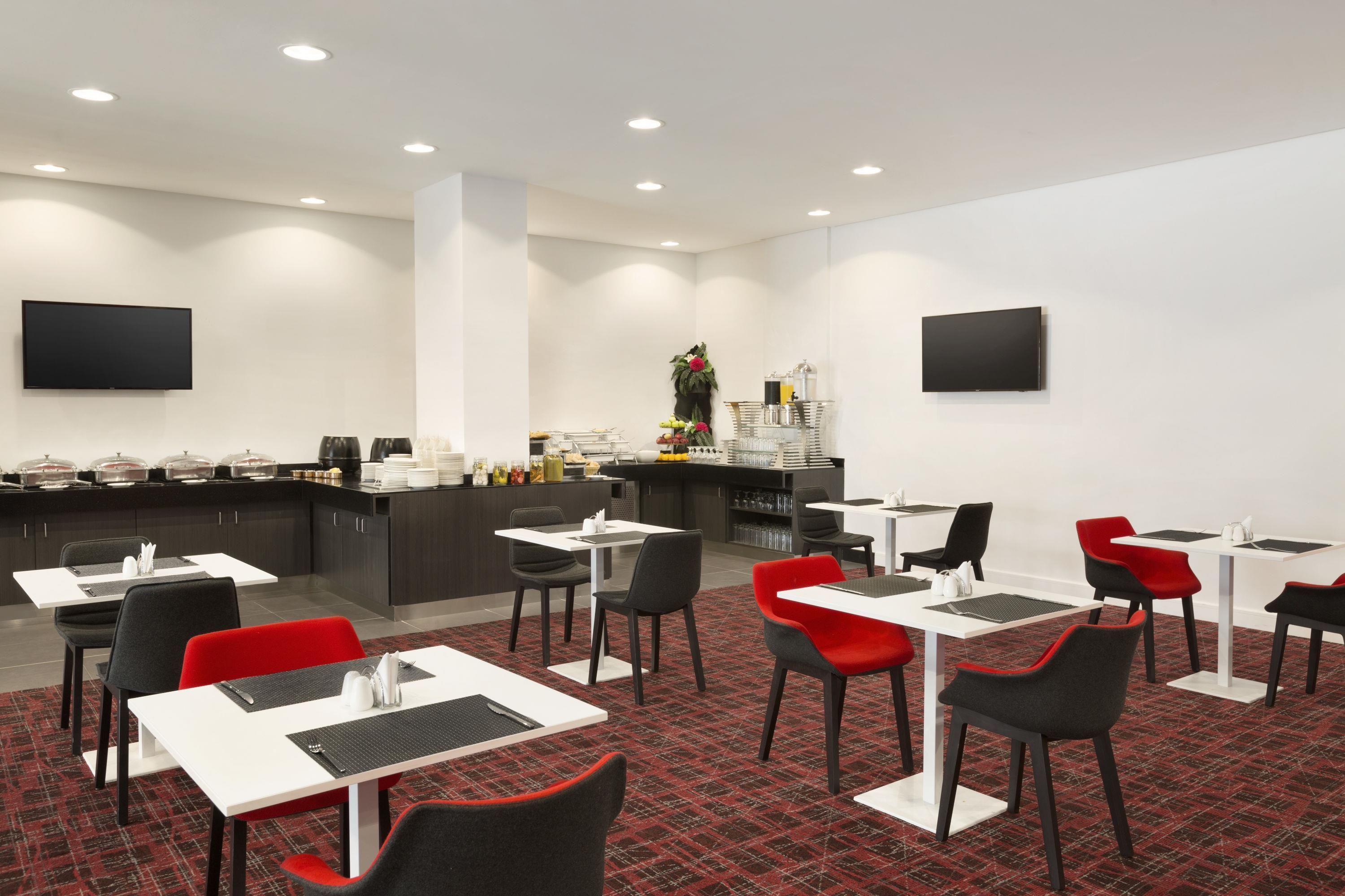 Un espace repas moderne avec des tables et des chaises, un coin bar avec un comptoir et des étagères, et deux téléviseurs à écran plat fixés aux murs.