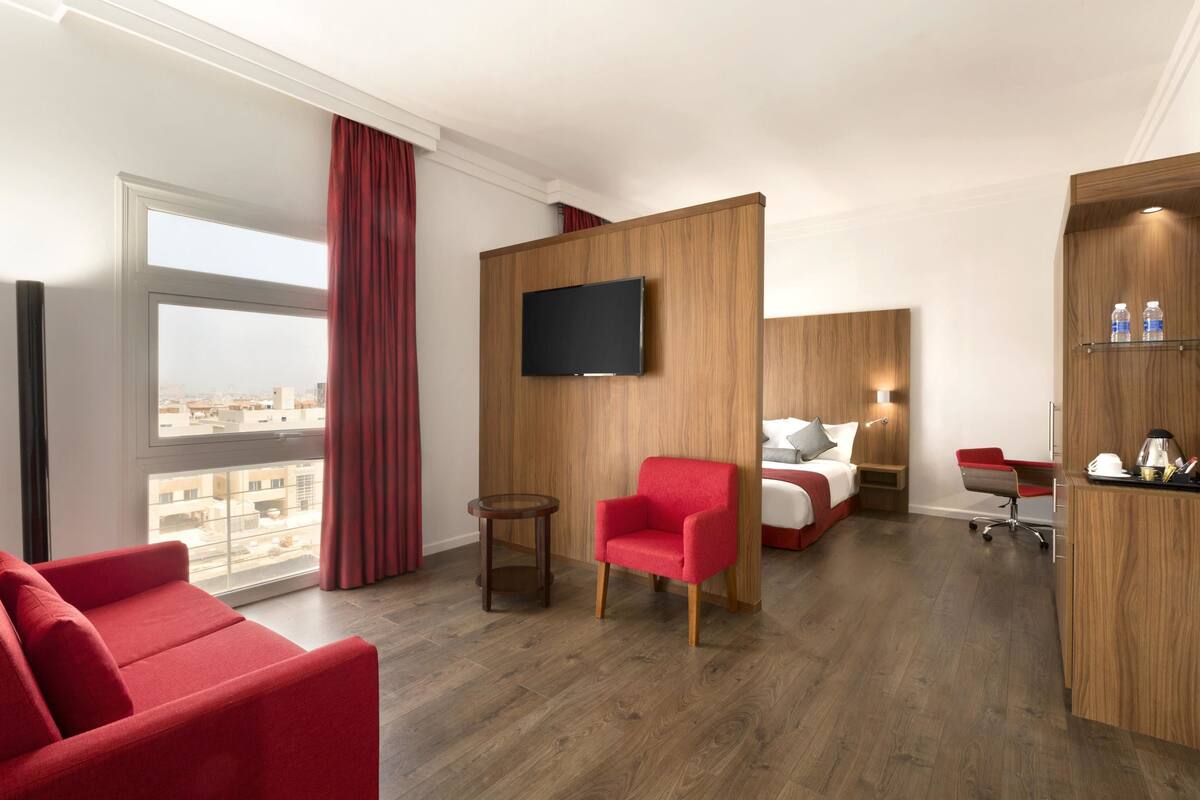 Ett hotellrum med ett stort fönster, en röd fåtölj, en säng med en tv och ett skrivbord med en stol.