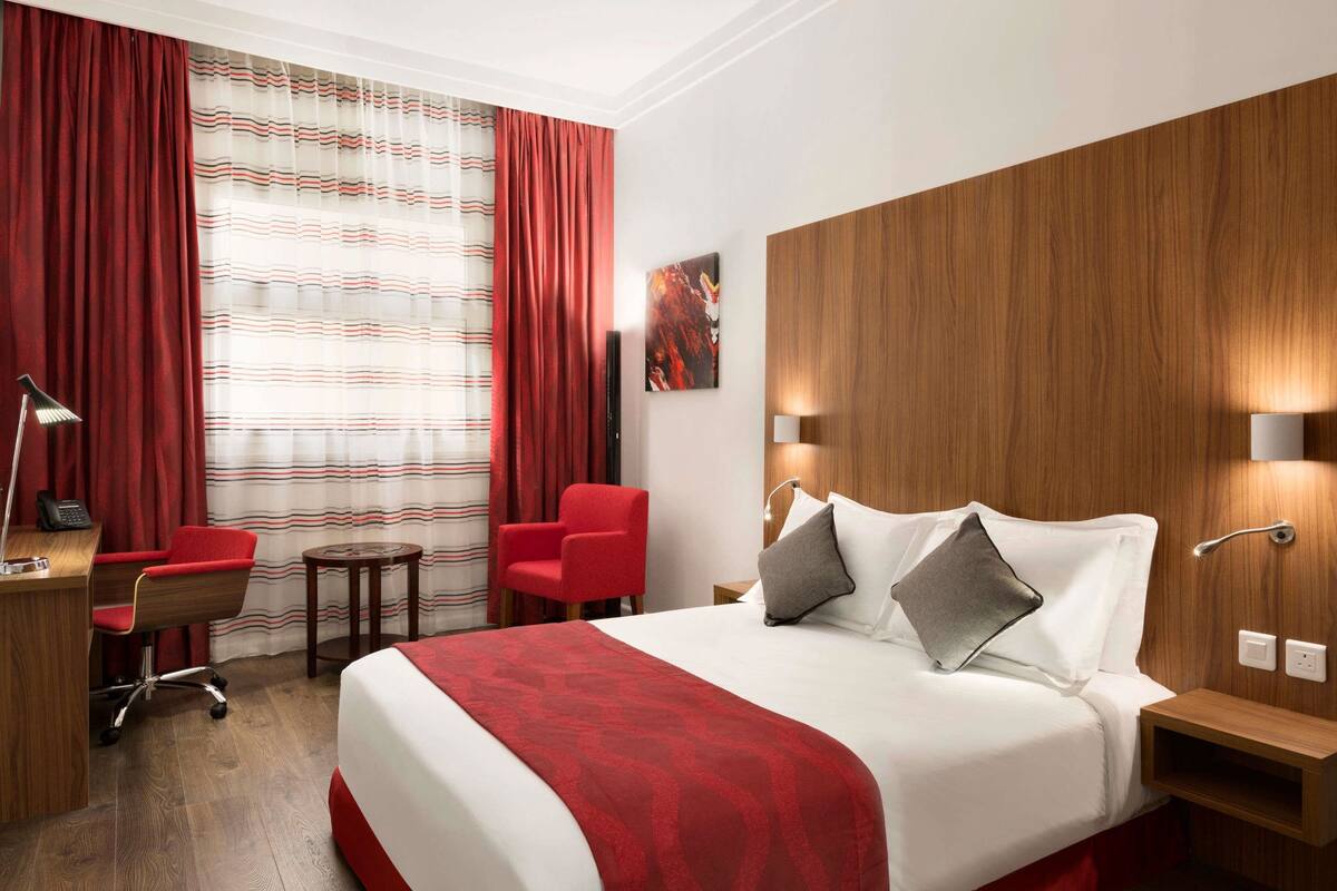 Ett hotellrum med en stor säng, ett skrivbord med en stol, ett litet bord och en röd fåtölj.