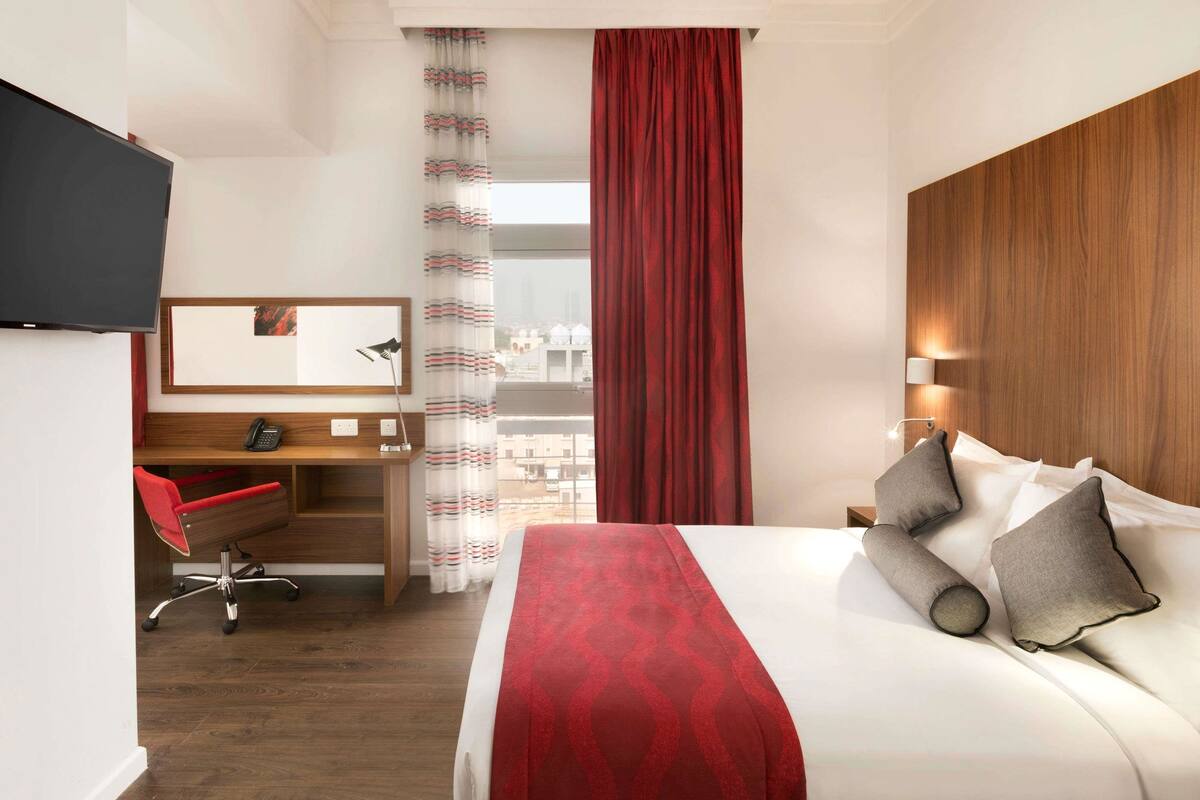 Ett hotellrum med en säng, ett skrivbord, en stol, en tv och ett fönster med gardiner.