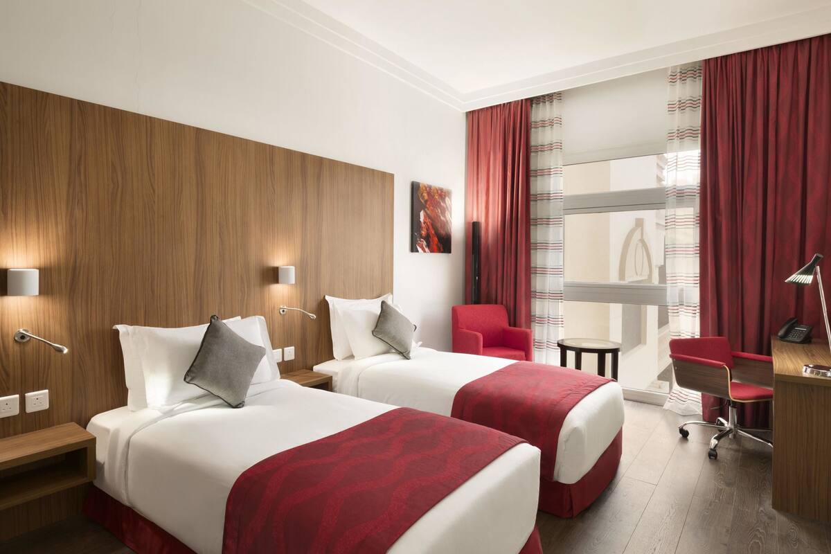 Ett hotellrum med två sängar, ett skrivbord, en stol och ett stort fönster med gardiner.