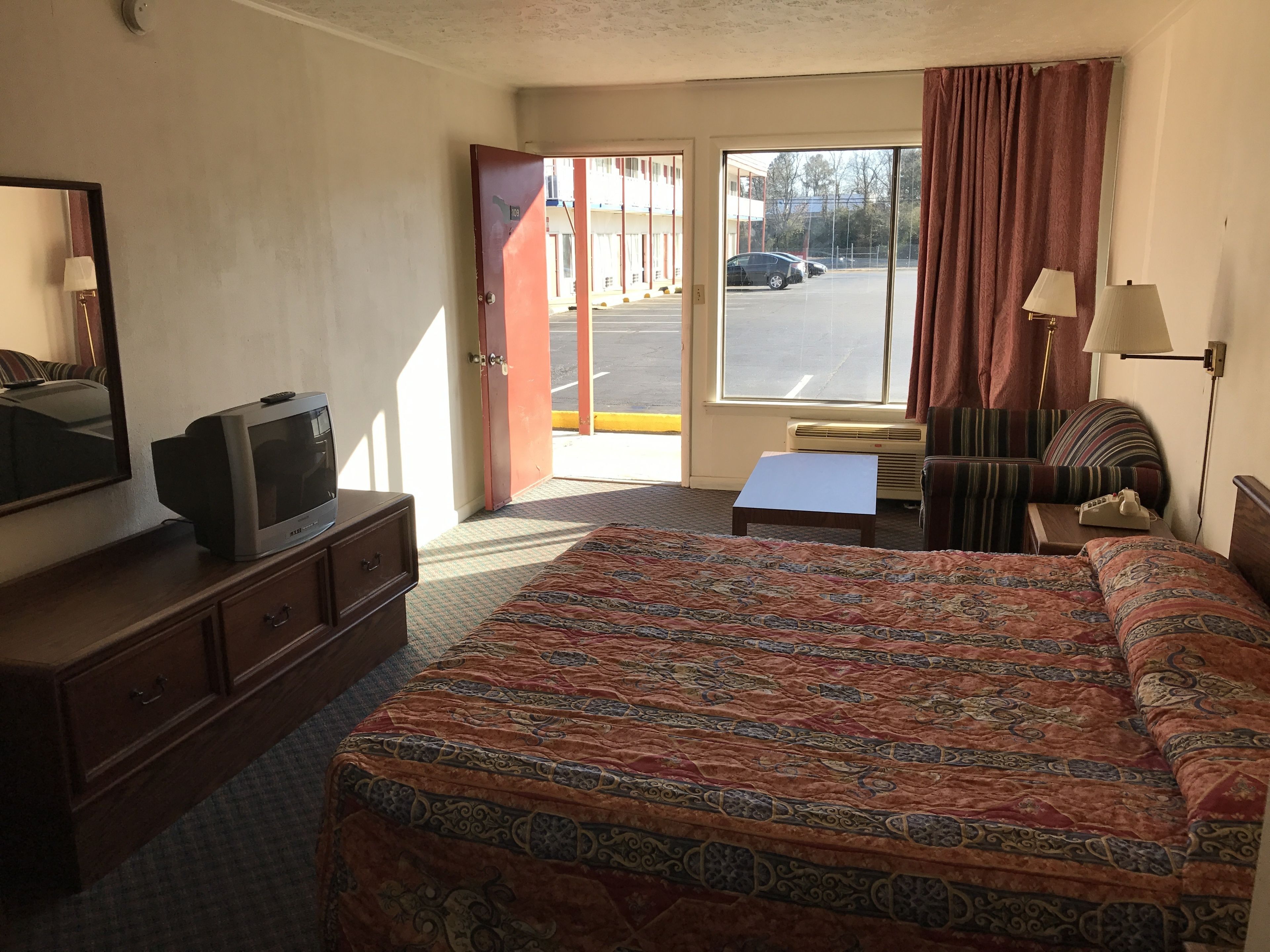 A hotel room with a bed, a dresser, a television, a window with curtains, and a view of the parking lot.