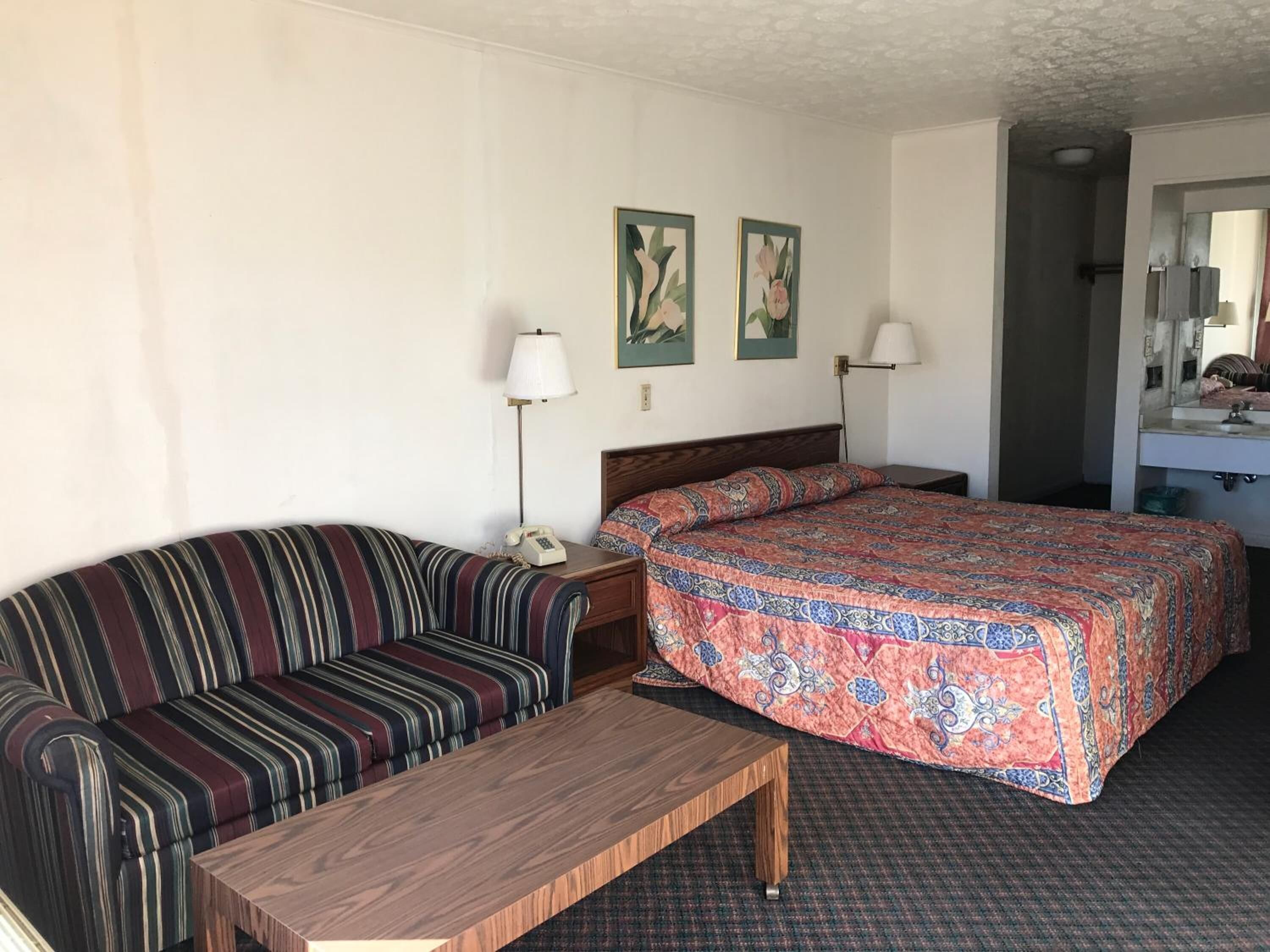 A hotel room with a striped sofa, a wooden coffee table, a bed with a patterned bedspread, two wall-mounted lamps, and a mirror above the sink.