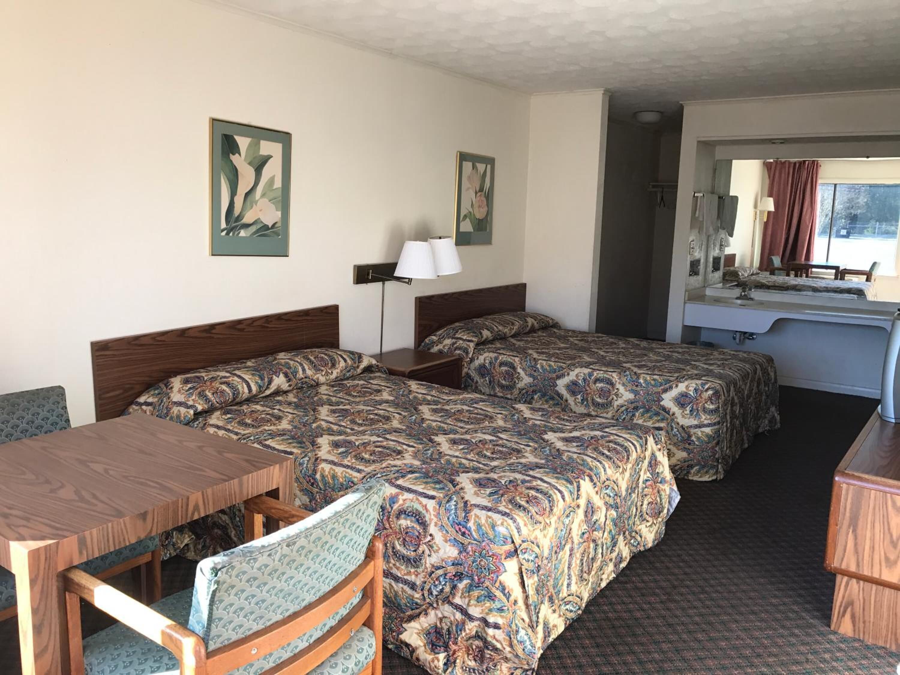 Habitación de hotel con dos camas, un escritorio, una silla, una lámpara y un baño visible a través de una puerta abierta.