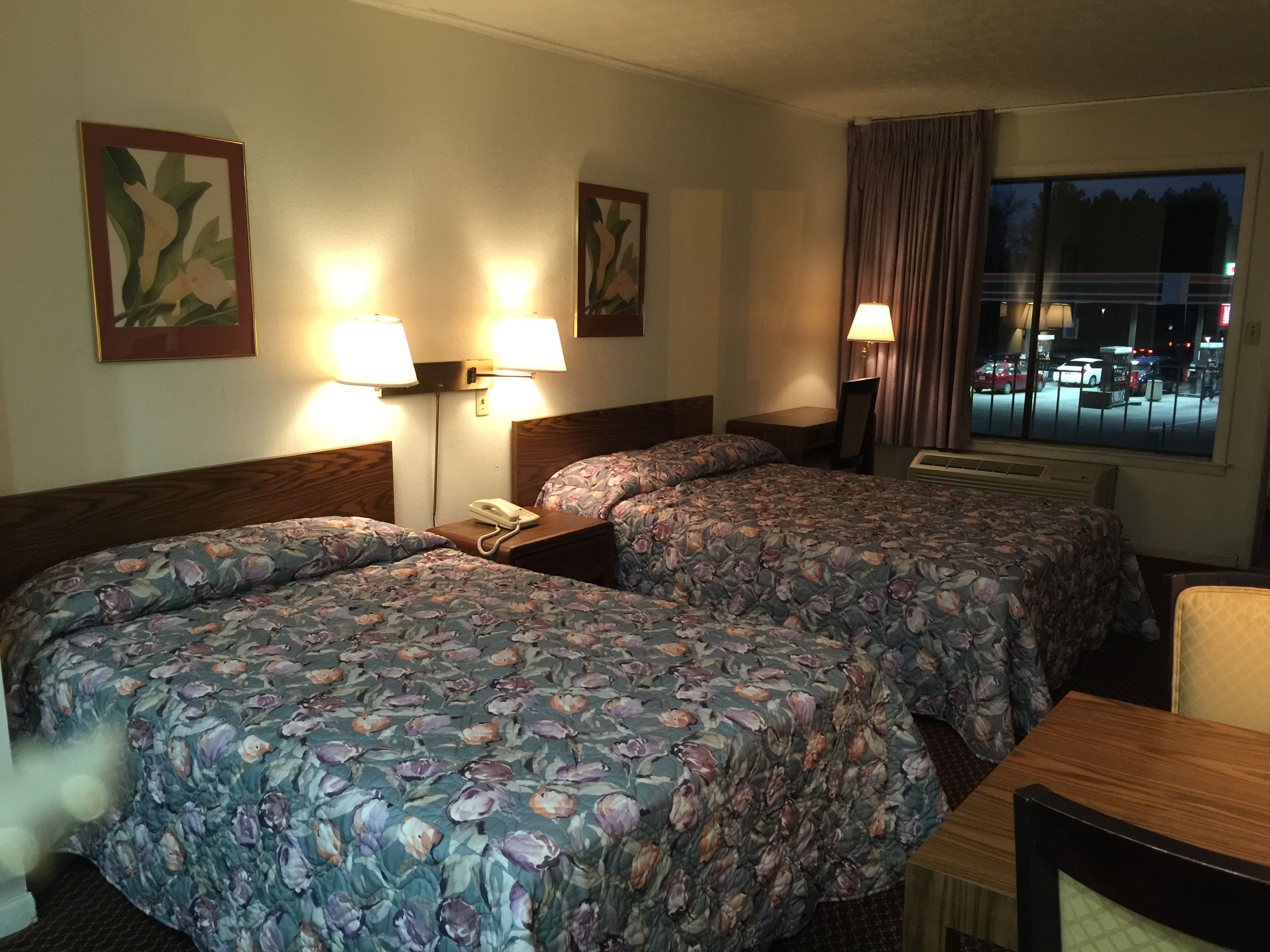 A hotel room with two beds, each with floral bedding, bedside lamps, and a nightstand with a phone. There is a window with a view of the city at night.